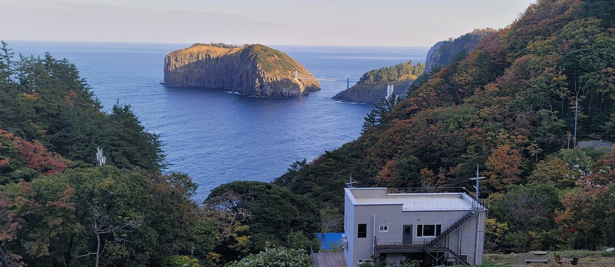 A scenic view showcases a modest building nestled within lush greenery, overlooking a vast ocean. In the distance, an island is visible, framed by gentle waves and rocky cliffs. The landscape is accentuated by autumn colors, creating a serene coastal atmosphere.