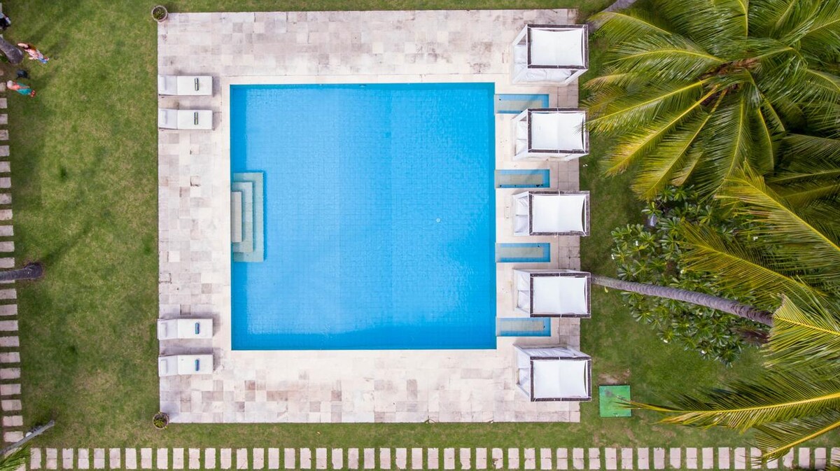 An aerial view reveals a tranquil pool surrounded by lush greenery and well-manicured grass. The pool area features several white lounge beds and a spacious deck, enhancing the relaxing atmosphere. Bright green palm trees frame the scene, contributing to the tropical setting.