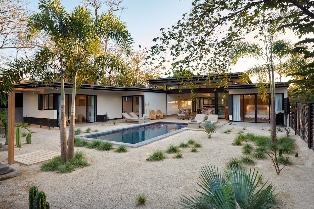 A modern villa is surrounded by landscaped gardens featuring palm trees and tropical plants. The spacious private pool is centrally located, reflecting the sunlight. Large glass doors connect the indoor living area with the outdoor space, creating a seamless flow for relaxation and enjoyment.