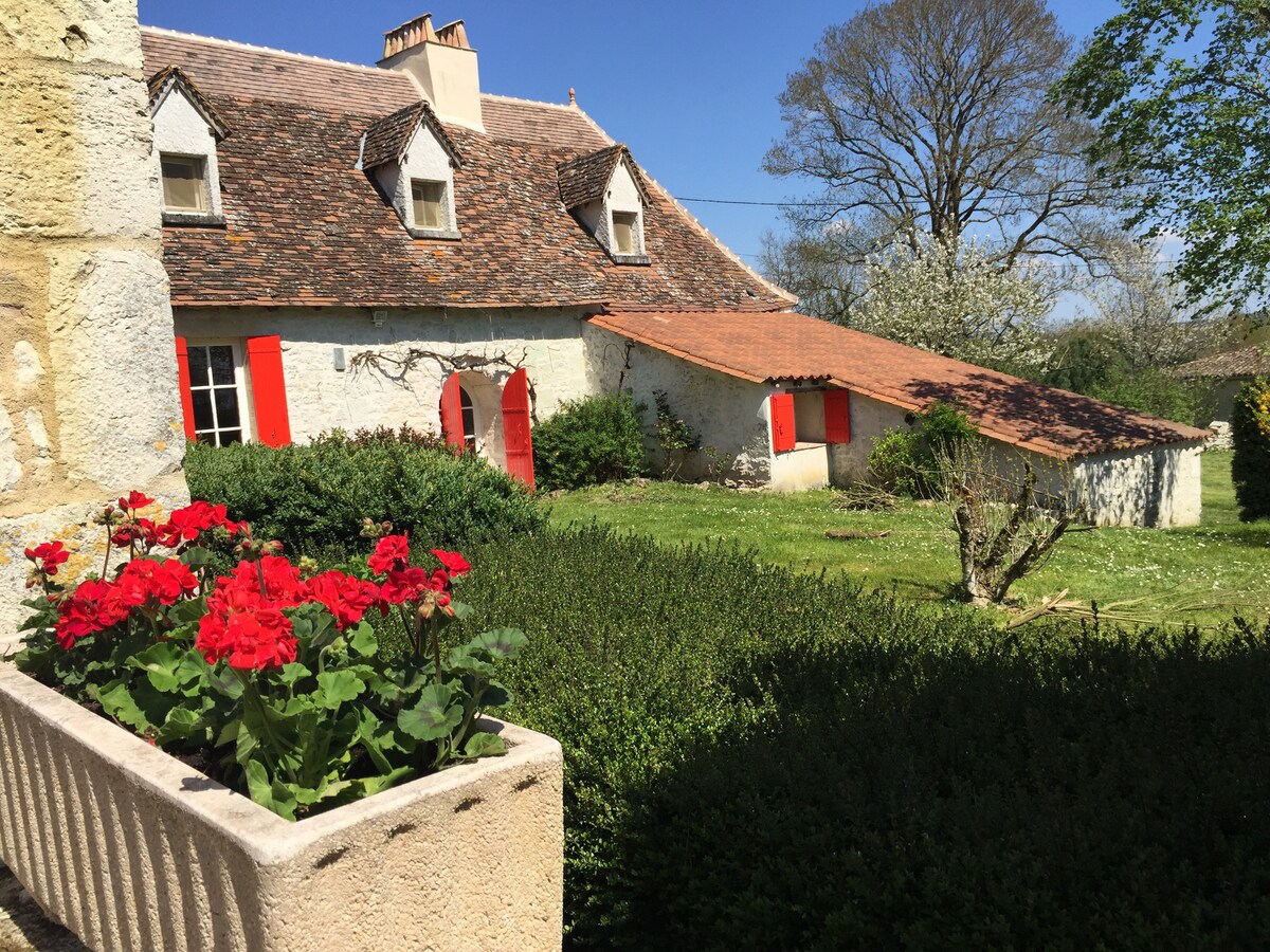 A charming exterior of a stone home is shown, featuring red-shuttered windows and a rustic roof. Vibrant red flowers in a planter add a touch of color, while a lush green lawn and trees create a serene environment.