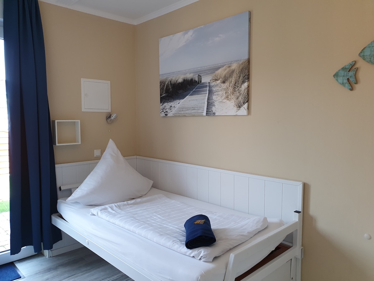 A minimalist sleeping area is equipped with a spacious bed, adorned with a white comforter and a large pillow positioned at an angle. A coastal-themed piece of wall art decorates the beige wall, while natural light enters through a glass door, complemented by dark blue curtains.