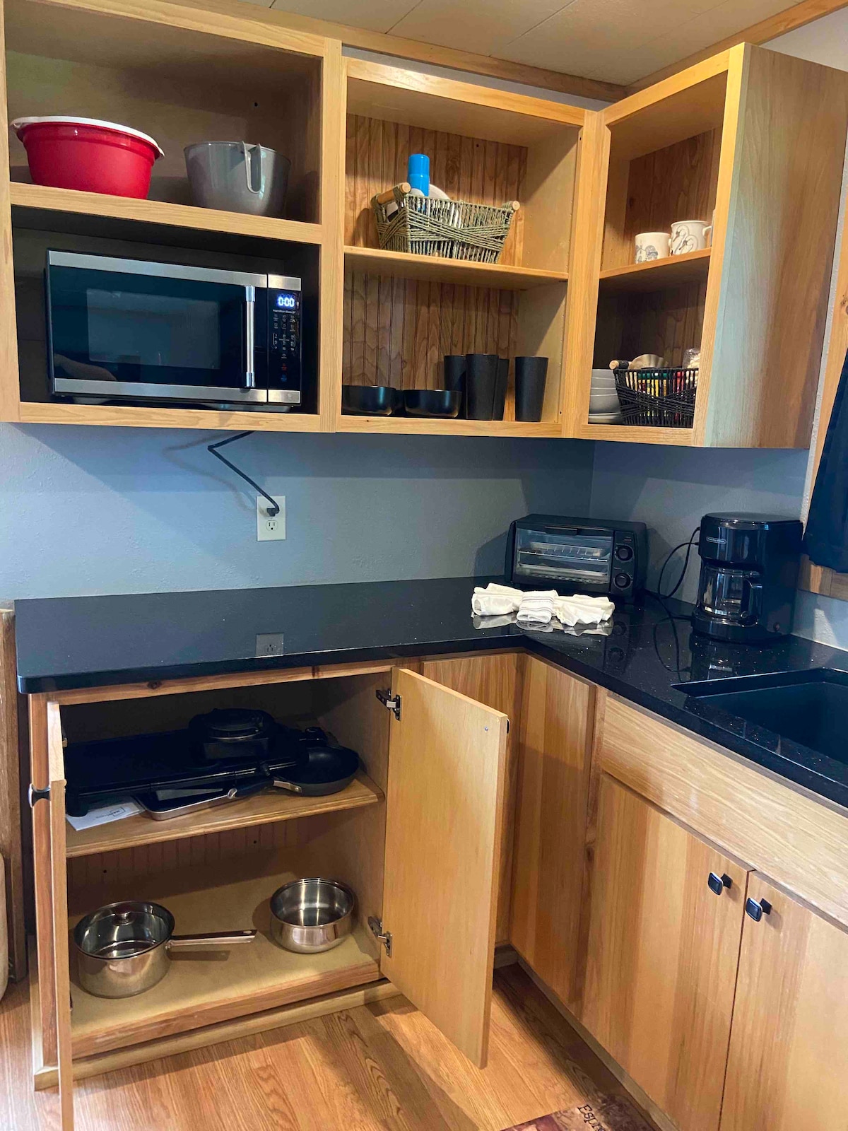 A functional kitchen area is displayed, featuring wooden cabinetry and a black countertop. A microwave and coffee maker are prominently positioned on the counter. Below, a cabinet is open, revealing metal cookware and a toaster oven. Organized shelving above contains various kitchen supplies.