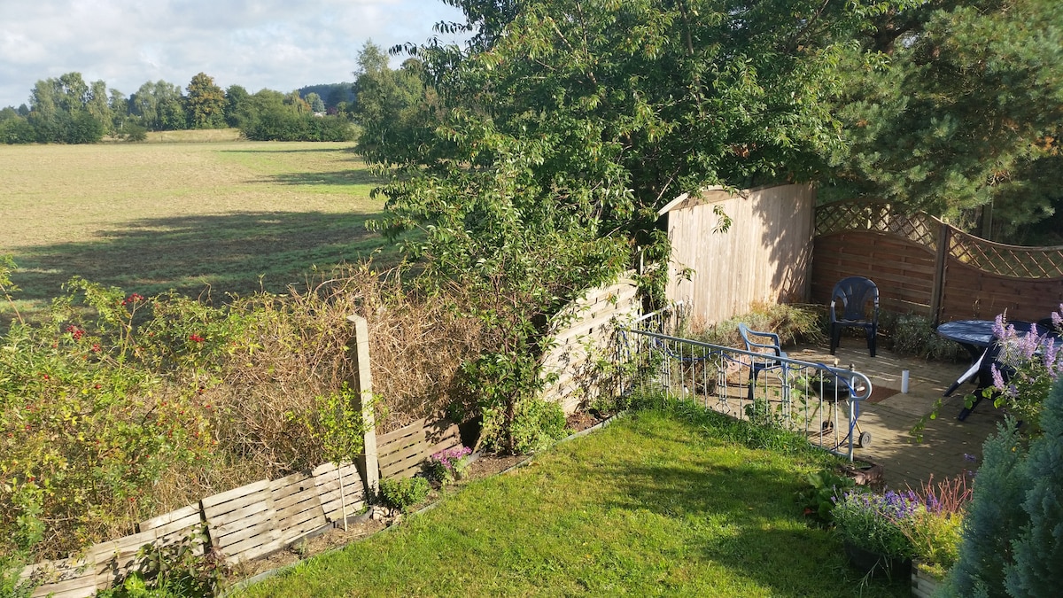 A well-maintained outdoor space is situated at the property's rear, featuring a neat lawn bordered by shrubs and flowers. A small patio area includes a metal table and chairs, providing an ideal view of the expansive field beyond, framed by trees and distant hills.