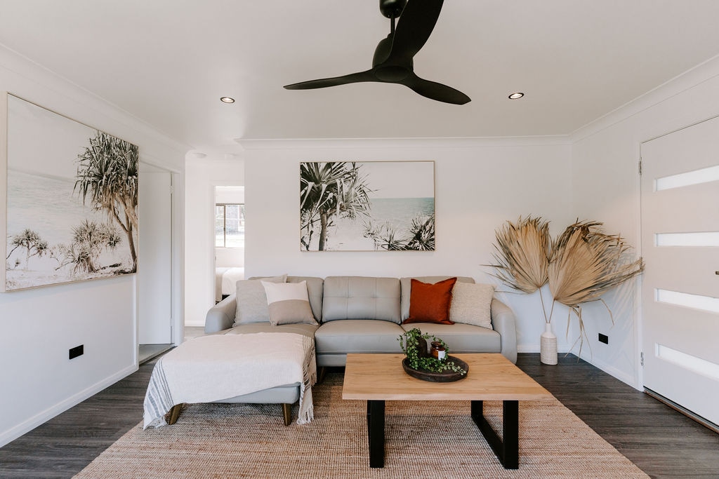 A contemporary living area features a light-colored sectional sofa with a textured throw and a decorative pillow. A wooden coffee table sits at the center, adorned with a small plant and decorative items. Two large artworks depicting coastal scenes adorn the walls, while a ceiling fan enhances airflow.