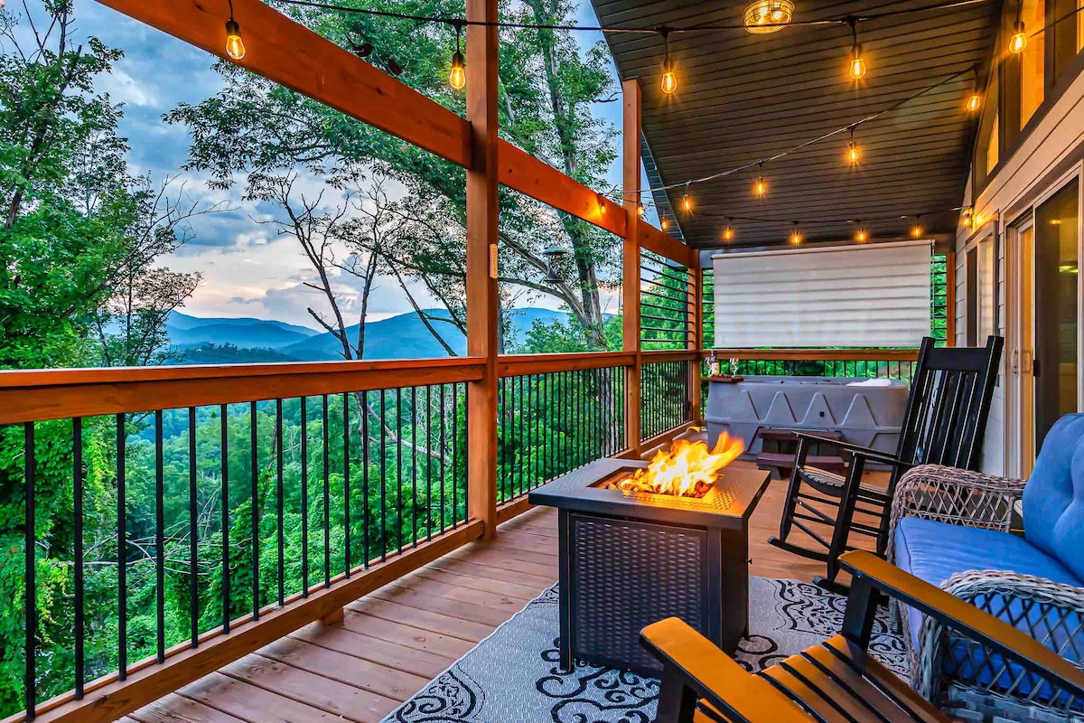 A spacious covered deck offers stunning mountain views, highlighted by string lights creating a warm ambiance. Comfortable seating includes rocking chairs and a blue cushioned chair surrounding a fire pit, perfect for cozy gatherings. Lush greenery and distant mountains enhance the serene atmosphere.
