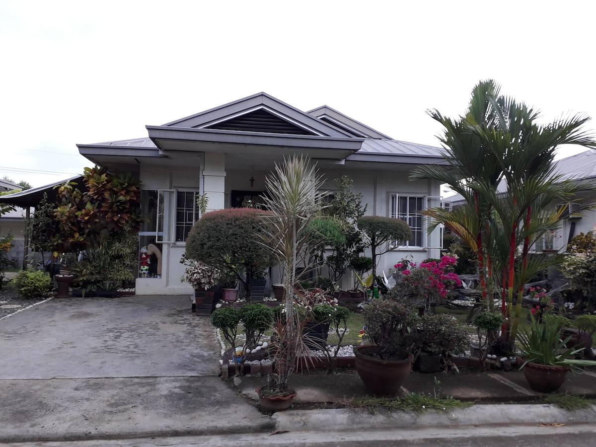 The front view of the property displays a single-story home with a peaked roof, surrounded by well-manicured gardens featuring various plants and flowers. A paved pathway extends to the entrance, leading to a welcoming porch area. Parking space is visible in the front.