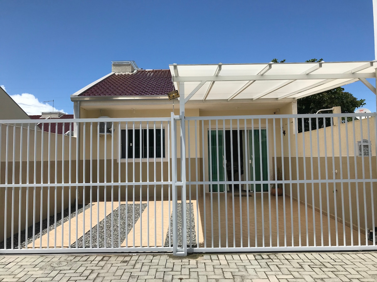 A newly constructed house is enclosed by a white gate and features a covered porch area. The exterior shows a combination of tiled and gravel surfaces, accompanied by large glass doors that lead into the house. A clear blue sky serves as the backdrop.