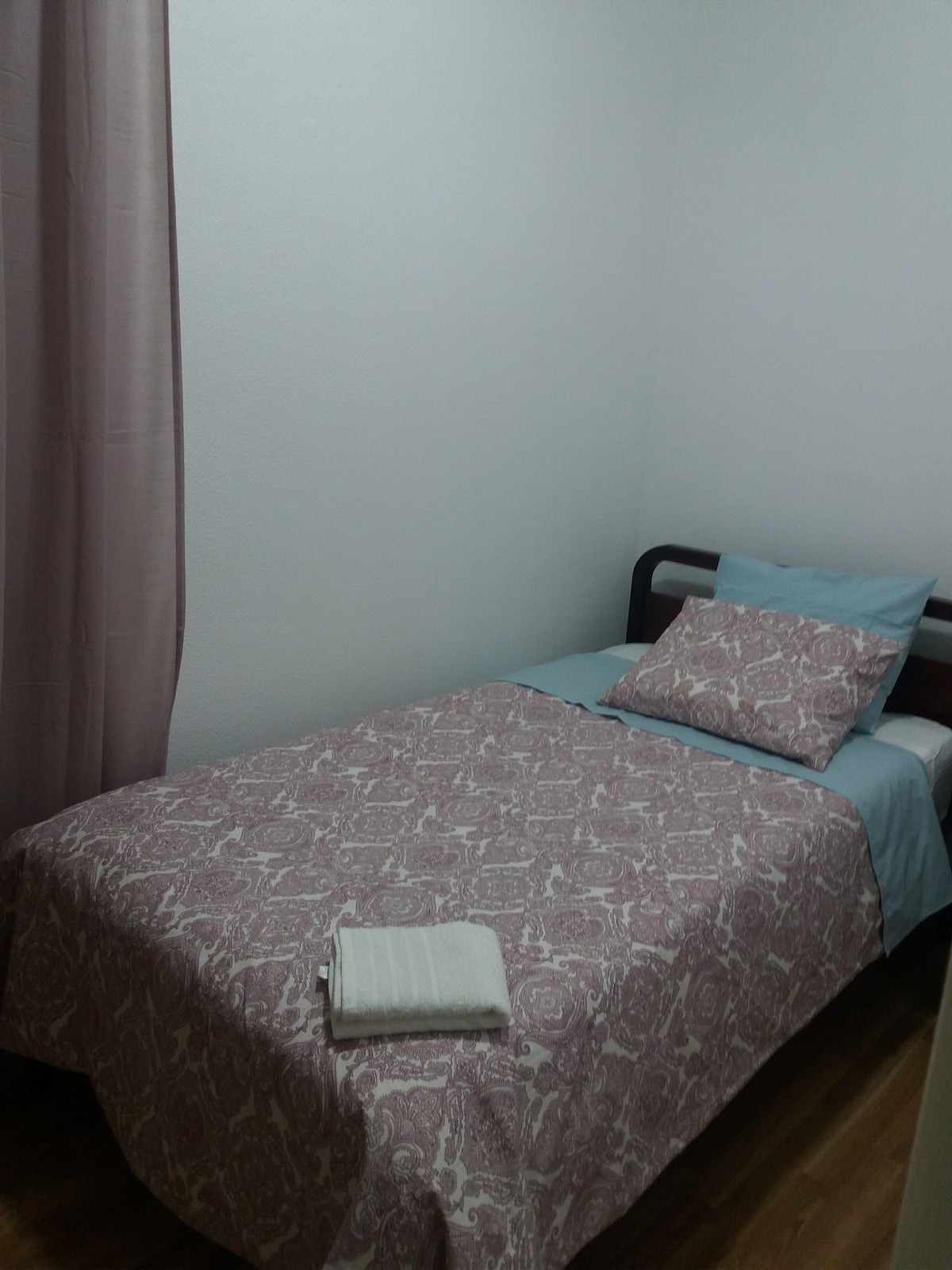 A single bed is arranged with a patterned bedspread and decorative pillow. A light blue sheet complements the bedding. A neatly folded towel is placed at the foot of the bed. Soft natural light filters through a nearby window covered by a light curtain.