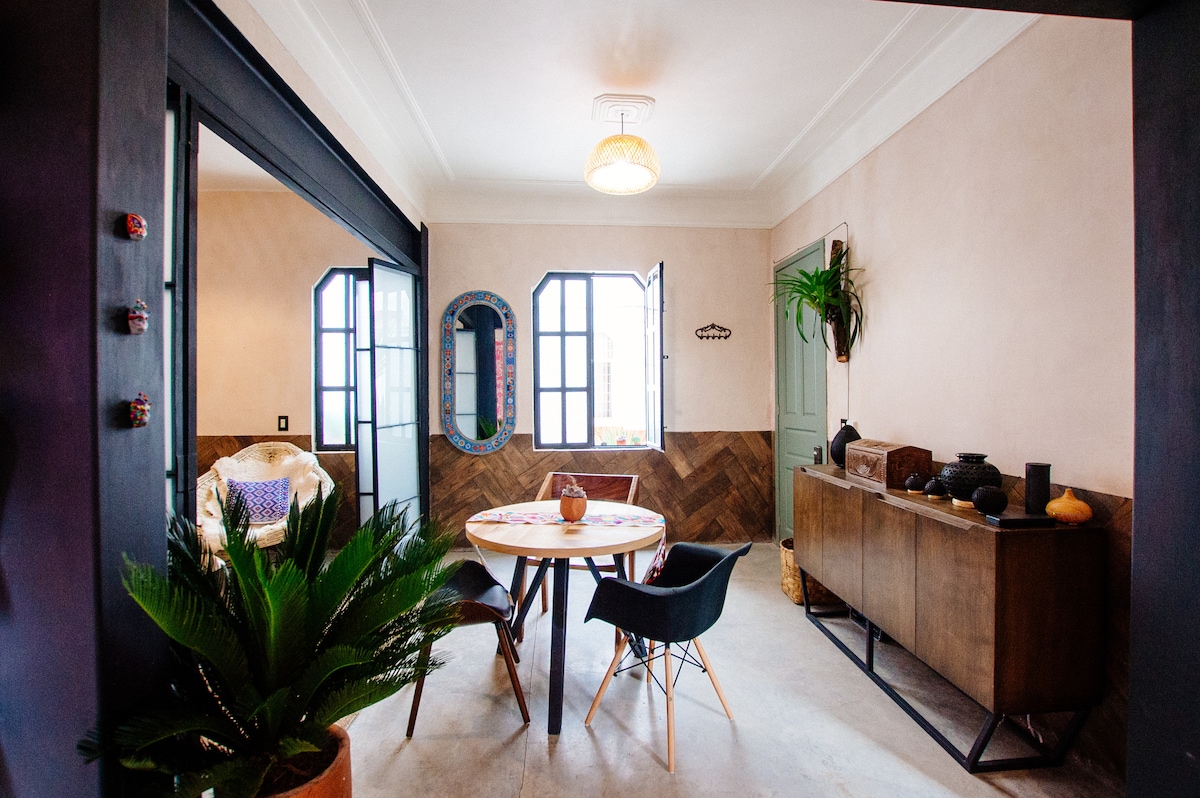 A stylish dining area is presented with a round wooden table surrounded by black chairs. Two large windows allow natural light to flow in, while a green plant adds a touch of nature. The room features beige walls and a charming wooden sideboard, enhancing the inviting atmosphere.