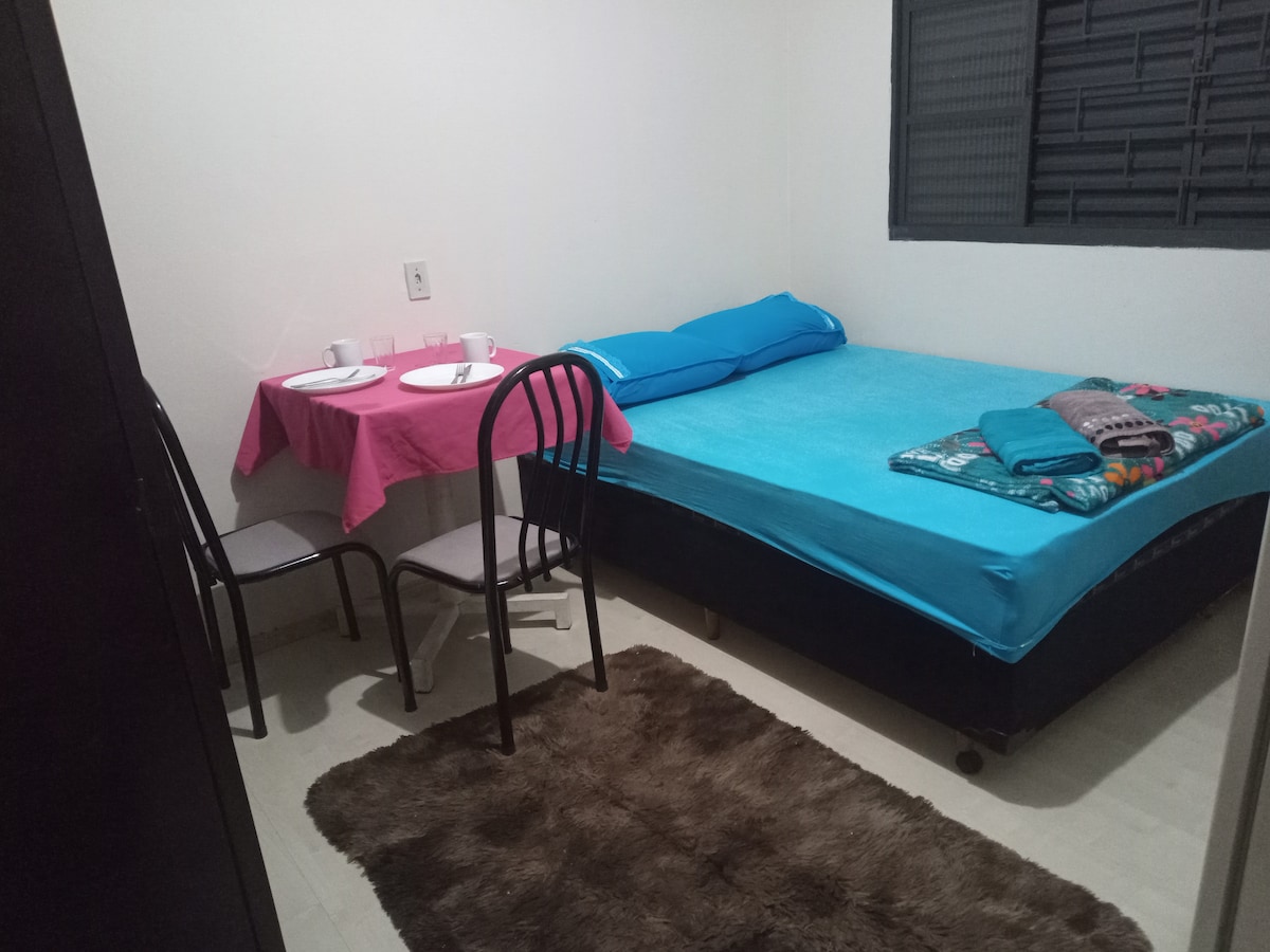 A simple room is furnished with a spacious bed covered in bright blue bedding. A small table set for two is placed nearby, adorned with white plates and cups. A cozy rug is positioned on the floor, complementing the overall decor. A window offers natural light.