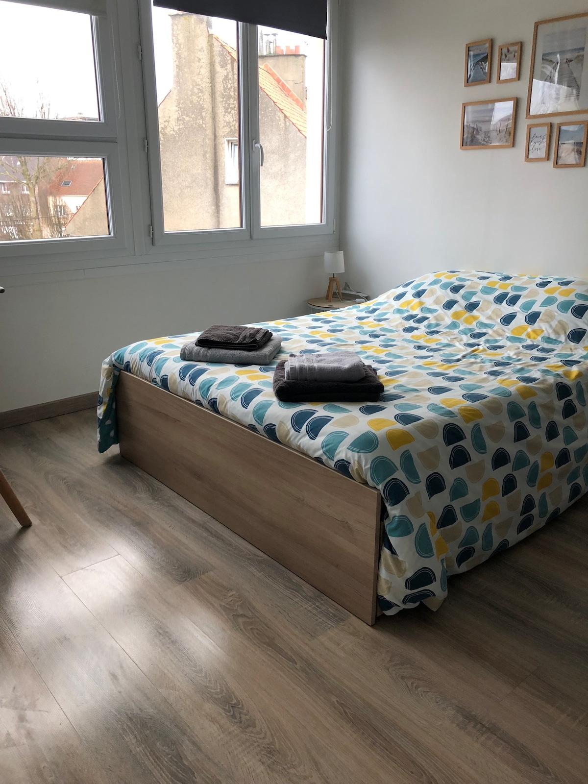 A spacious bedroom features a bed with a colorful patterned coverlet. Two neatly folded towels rest on the bed. Natural light filters in through multiple windows, illuminating the wooden floor and enhancing the room's inviting atmosphere.