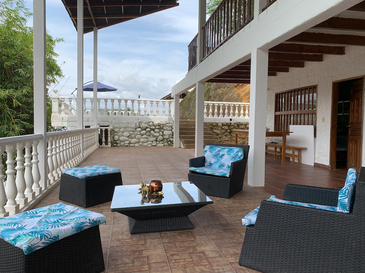 An outdoor seating area features a modern coffee table surrounded by four comfortable chairs with tropical-patterned cushions. The inviting space is shaded by a wooden overhang, and the surrounding architecture showcases a blend of stone and white columns, creating an airy atmosphere.