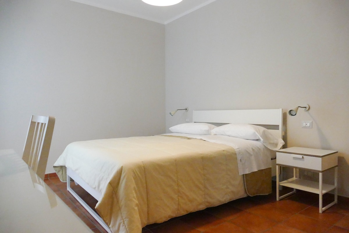 The room features a spacious bed dressed in neutral-colored linens, complemented by bedside tables with reading lamps on either side. A modern desk is positioned nearby, and natural light illuminates the space, highlighting the simple and clean design of the room.