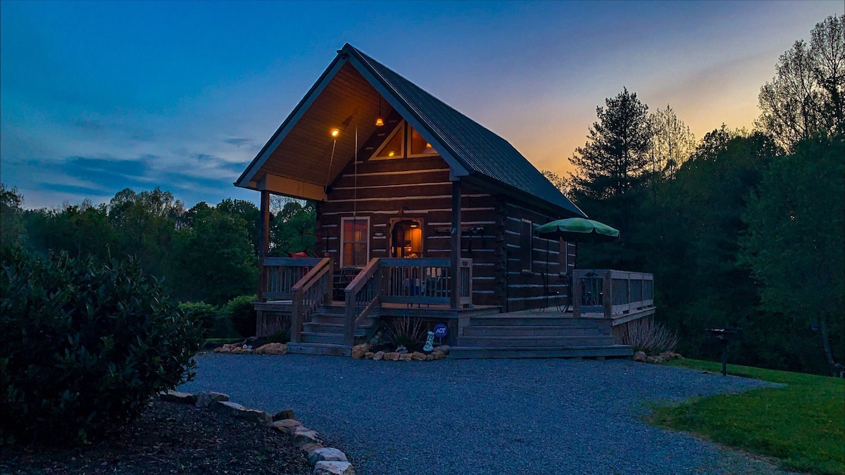 A charming log cabin is positioned on a hillside, featuring a welcoming porch illuminated by warm lighting. Surrounding greenery enhances the serene atmosphere, while a patio umbrella offers shade. The gravel driveway adds to the rustic appeal, with natural landscaping framing the entrance.