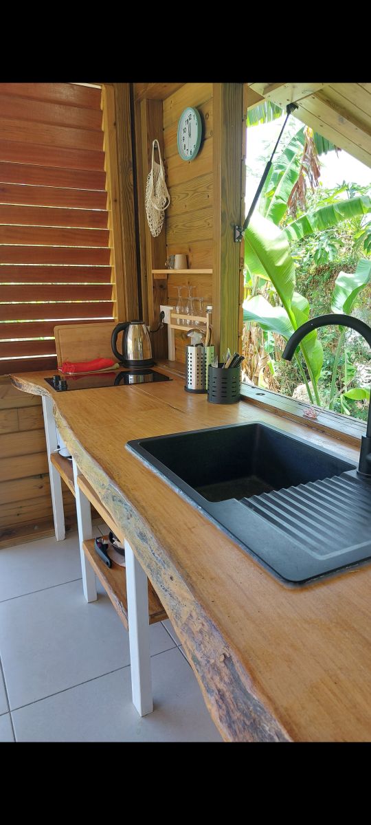 A wooden kitchen counter features a sleek black sink and kettle, complemented by organized utensils in a holder. Natural light filters through open wooden slats, highlighting green foliage visible outside, creating a connection with the environment.