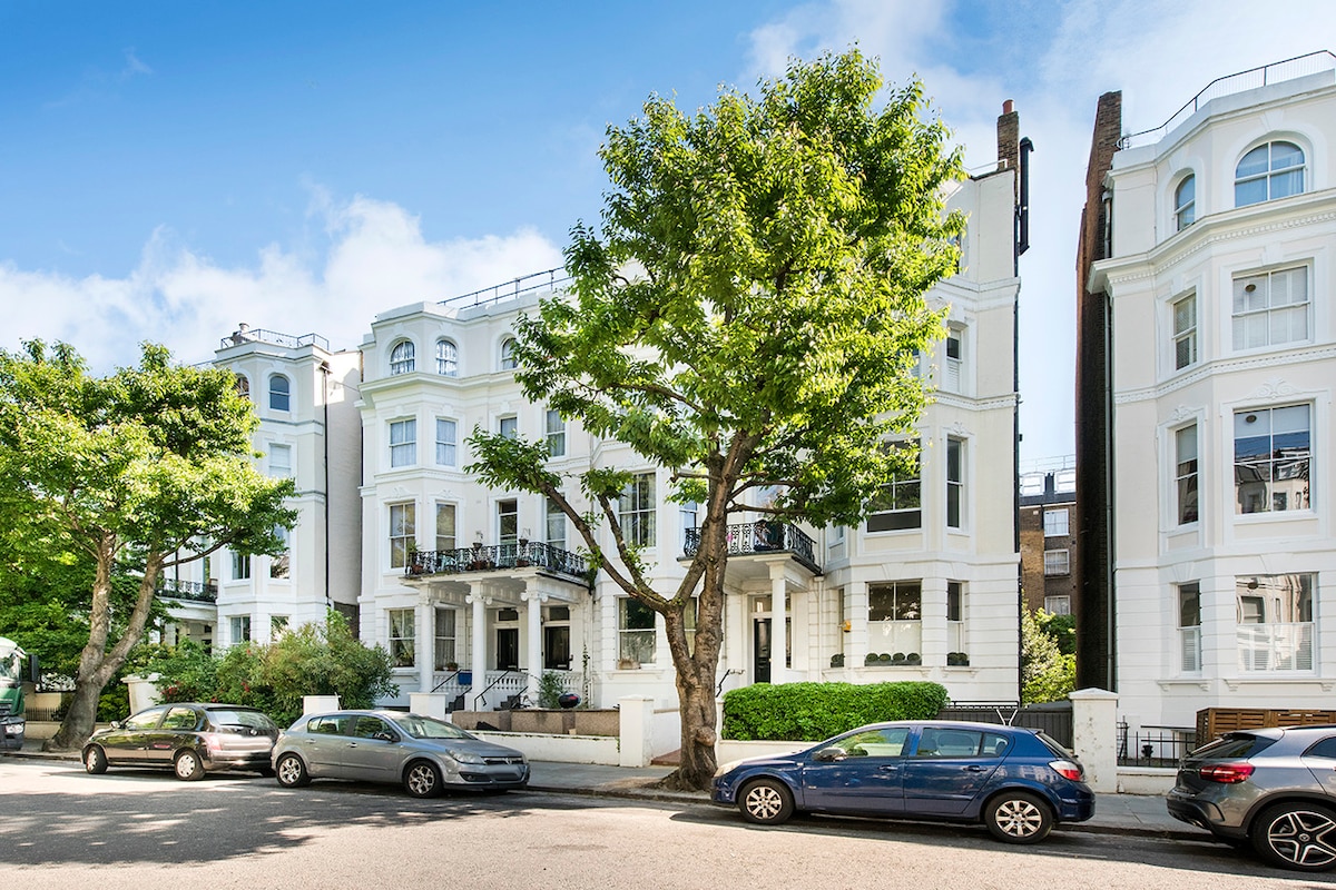 A row of elegant white buildings is visible, showcasing classic architecture with large windows and balconies. Lush green trees line the street, creating a calm atmosphere. Several parked cars are present along the quiet road, enhancing the residential charm of the neighborhood.