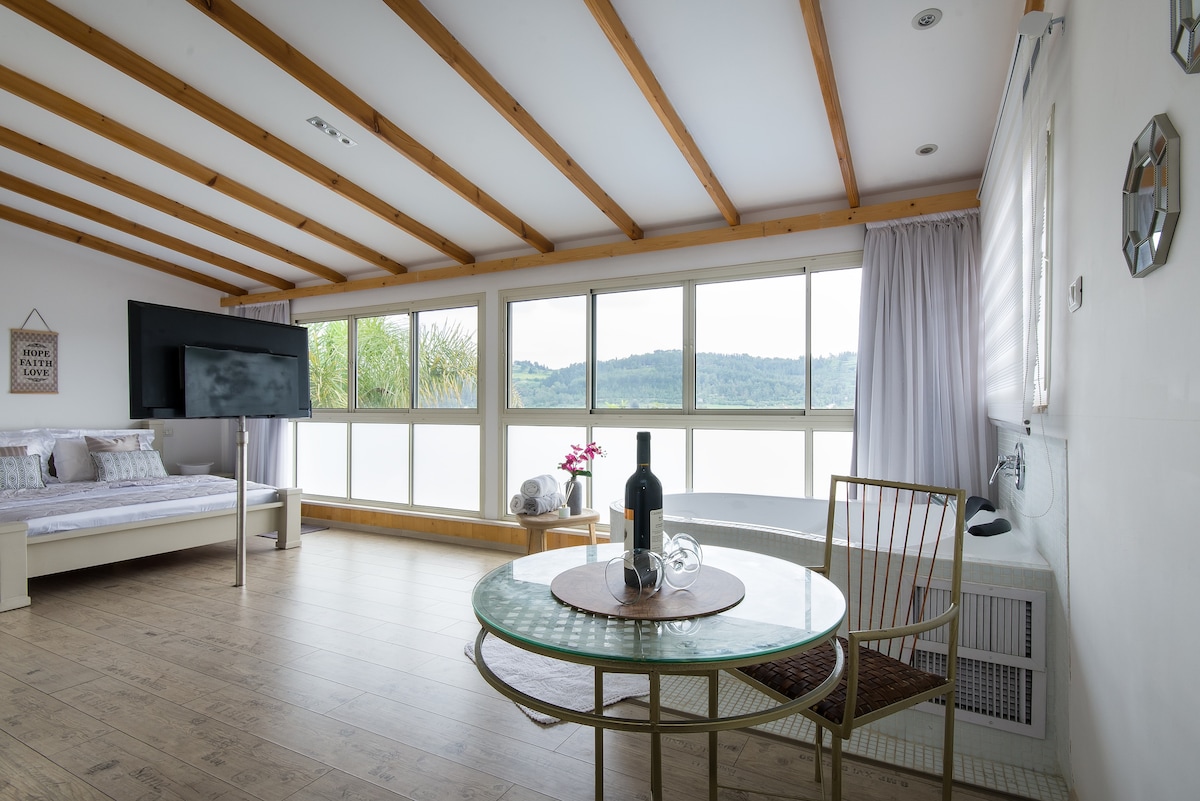 A spacious living area features large windows allowing for an abundance of natural light. A cozy bed is positioned near the windows, and a circular dining table with chairs is set in front of a flat-screen TV. Scenic views of the surrounding hills are visible.