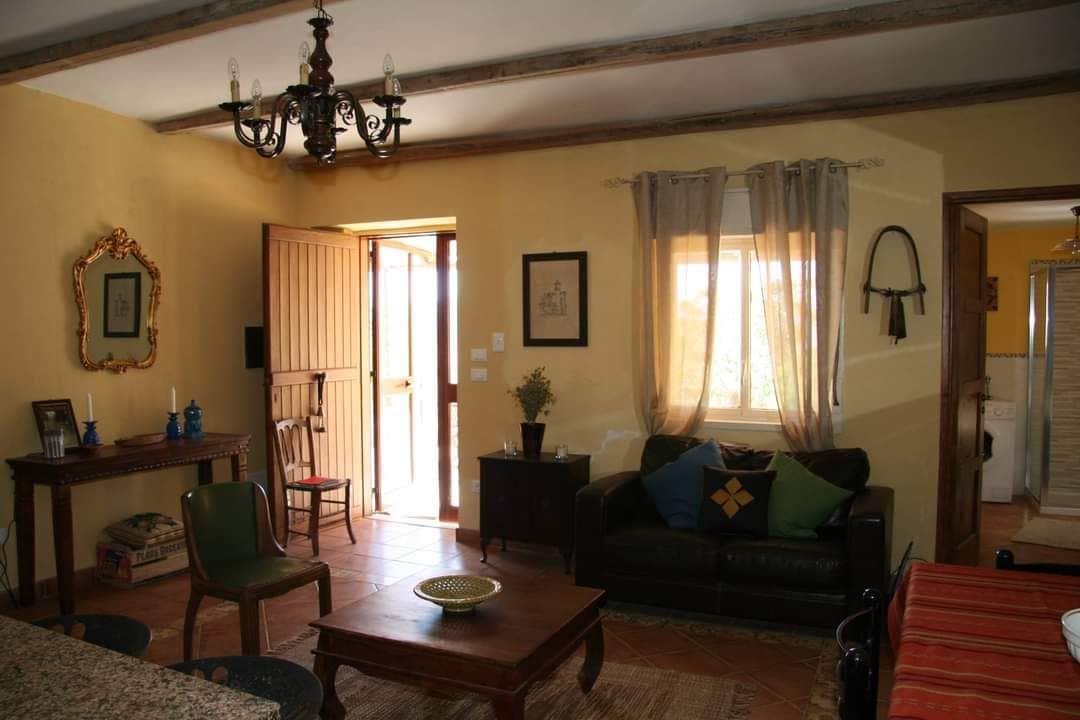 The inviting living area features warm yellow walls and exposed wooden beams. A comfortable black sofa is paired with a wooden coffee table. Natural light fills the space through the window and door, enhancing the cozy atmosphere. Decorative elements include a wall-mounted horse saddle and vintage artwork.