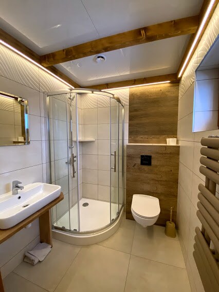 The spacious bathroom features a round glass shower enclosure, a modern sink with a wooden countertop, and a toilet. Wall panels combine white tiles with wooden accents, while soft LED lighting enhances the design. A towel rack and a small decorative element are also present.