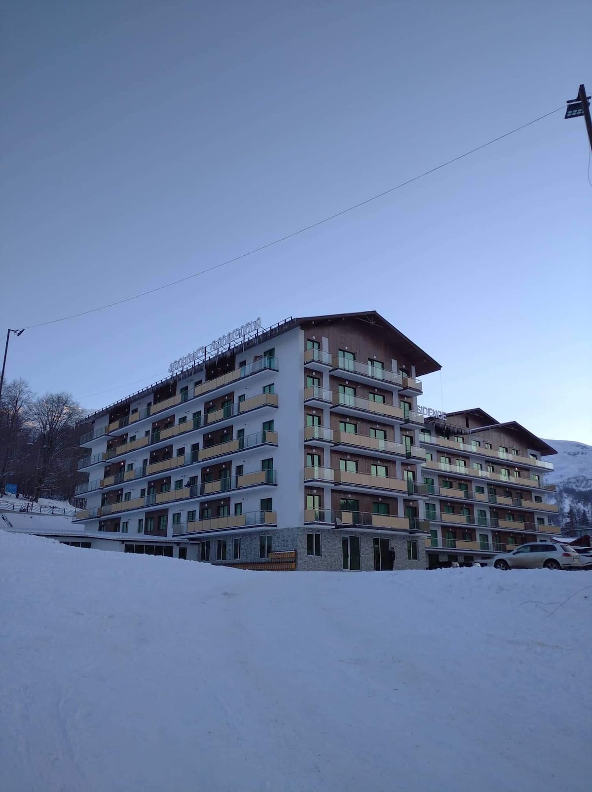 The multi-story building features a combination of light and dark exterior tones, with multiple balconies visible on each floor. Snow covers the ground around the structure, and the backdrop includes hillside terrain, indicating a serene environment.