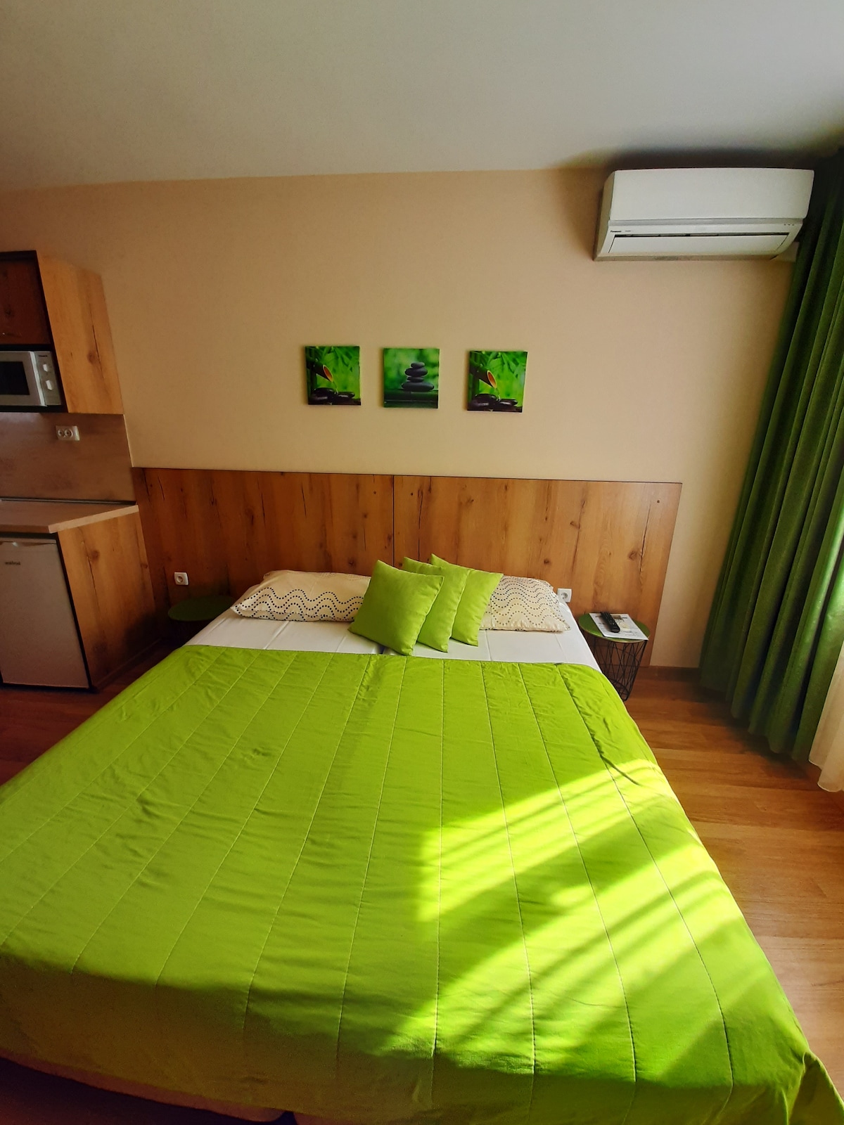 The studio features a spacious bed dressed in a bright green coverlet, accented by coordinating pillows. A minimalist wooden panel creates a warm backdrop, with three framed green artworks above the bed. Natural light filters through a nearby window, complemented by dark green curtains.