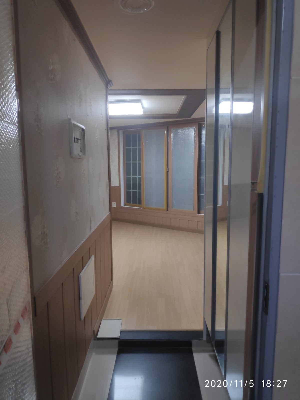 A hallway features light-colored wooden flooring and walls adorned with a subtle floral pattern. Large windows in the adjoining room allow natural light to fill the space. Bright overhead lighting illuminates the area, creating a welcoming atmosphere.