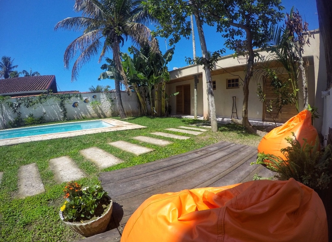 A sunny outdoor area features a turquoise swimming pool surrounded by well-maintained grass and tropical plants. Comfortable orange bean bags are positioned near a wooden deck, with a small flower pot adding a touch of color. The house is visible in the background, blending with the serene landscaping.