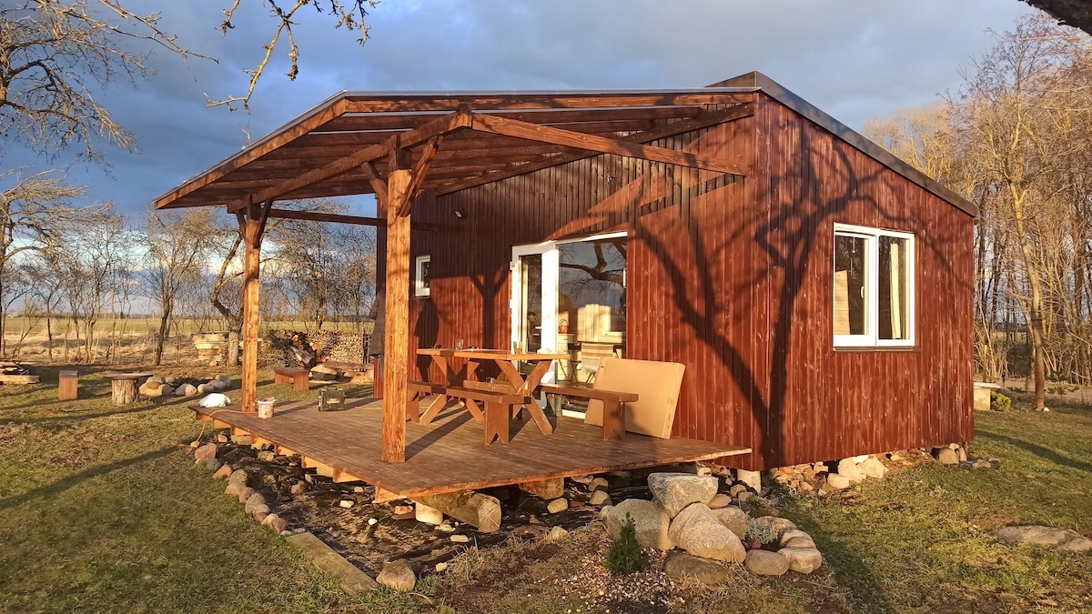 A wooden cabin with a covered porch is set against a serene landscape. The porch features a picnic table and seating area, with large windows providing natural light. Surrounding stones and greenery enhance the outdoor setting.