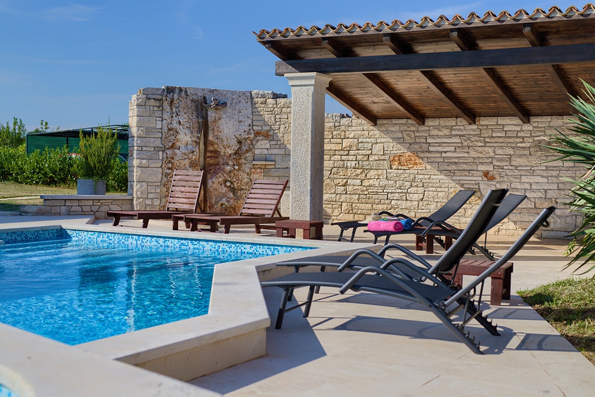 A swimming pool is surrounded by smooth stone tiles and shaded by a wooden pergola. Two lounge chairs are positioned beside the pool, offering a place to relax. The area is framed by greenery and features a rustic stone wall in the background.