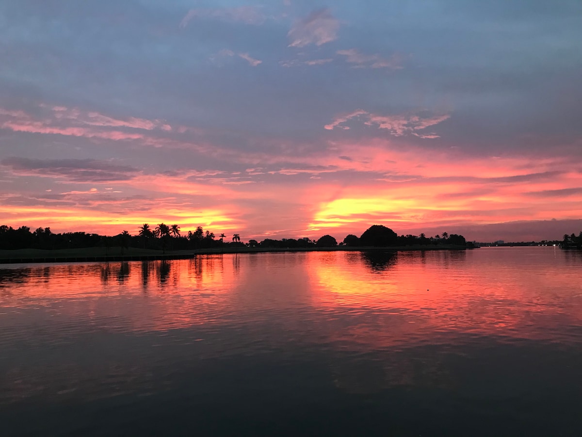 A serene waterfront scene captures a vibrant sunset, where the sky is painted in shades of pink and orange. The calm water reflects the colorful hues, with silhouettes of palm trees and distant islands adding depth to the landscape.