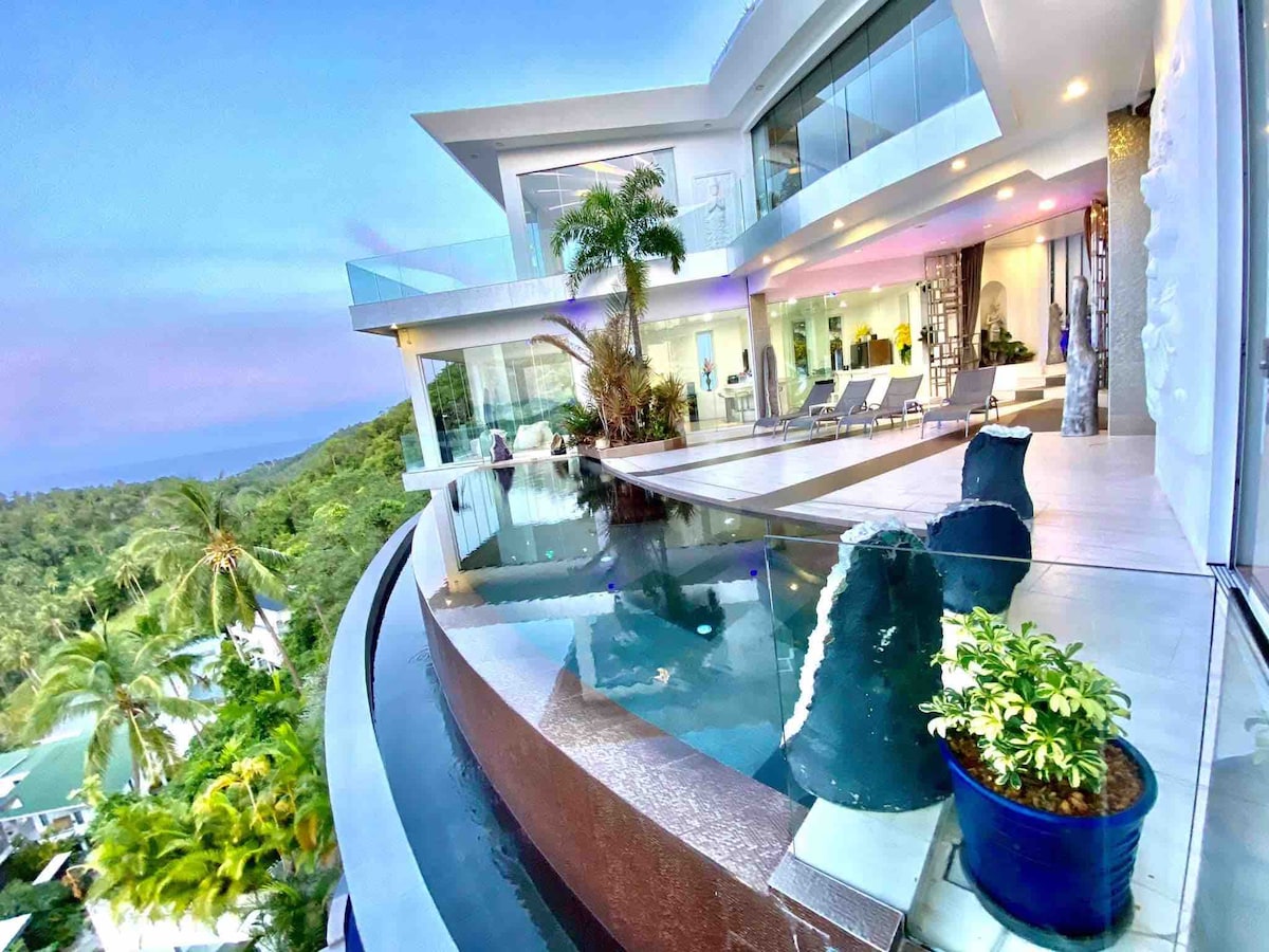 An infinity pool curves alongside a modern villa, offering stunning views of lush greenery. Lounge chairs are positioned for relaxation, while large glass doors invite natural light into the open living space. Tropical plants add a touch of nature to the elegant design.