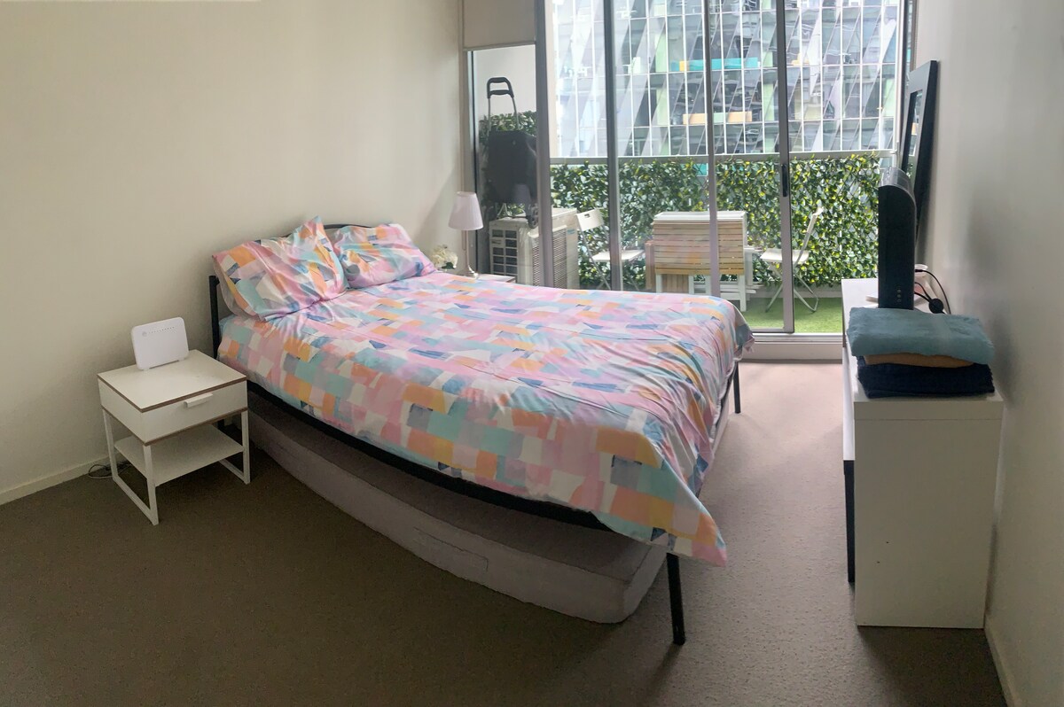 A cozy bedroom features a double-sized bed with a multicolored quilt and matching pillows. A bedside table with a lamp is positioned beside the bed. Large glass doors provide natural light and view of the outdoor greenery, complemented by a simple seating area outside.
