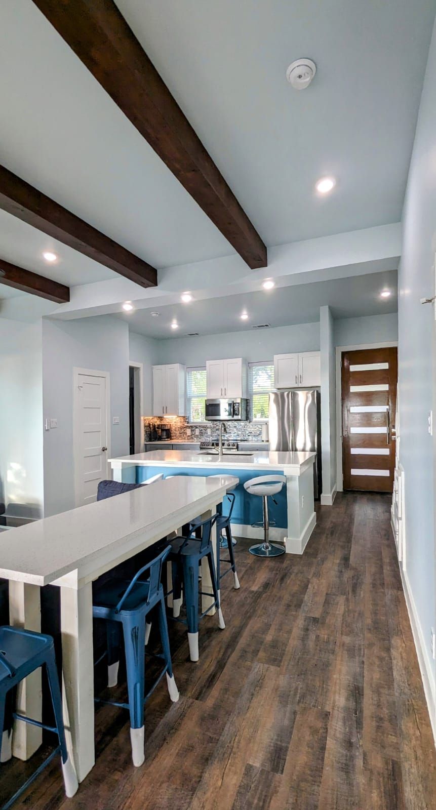 A modern kitchen features white cabinetry and stainless steel appliances, illuminated by recessed lighting. A long countertop provides seating with blue bar stools, complemented by wooden beams overhead. A door with clear glass panels adds to the overall open and airy feel.