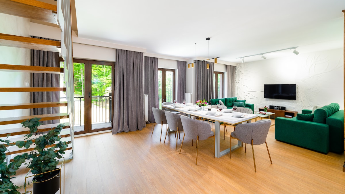 A modern living area features a large dining table set for a meal, accompanied by comfortable gray chairs. Two green sofas are arranged to encourage conversation, while large windows provide natural light and views of the surrounding greenery. Soft curtains frame the windows elegantly.