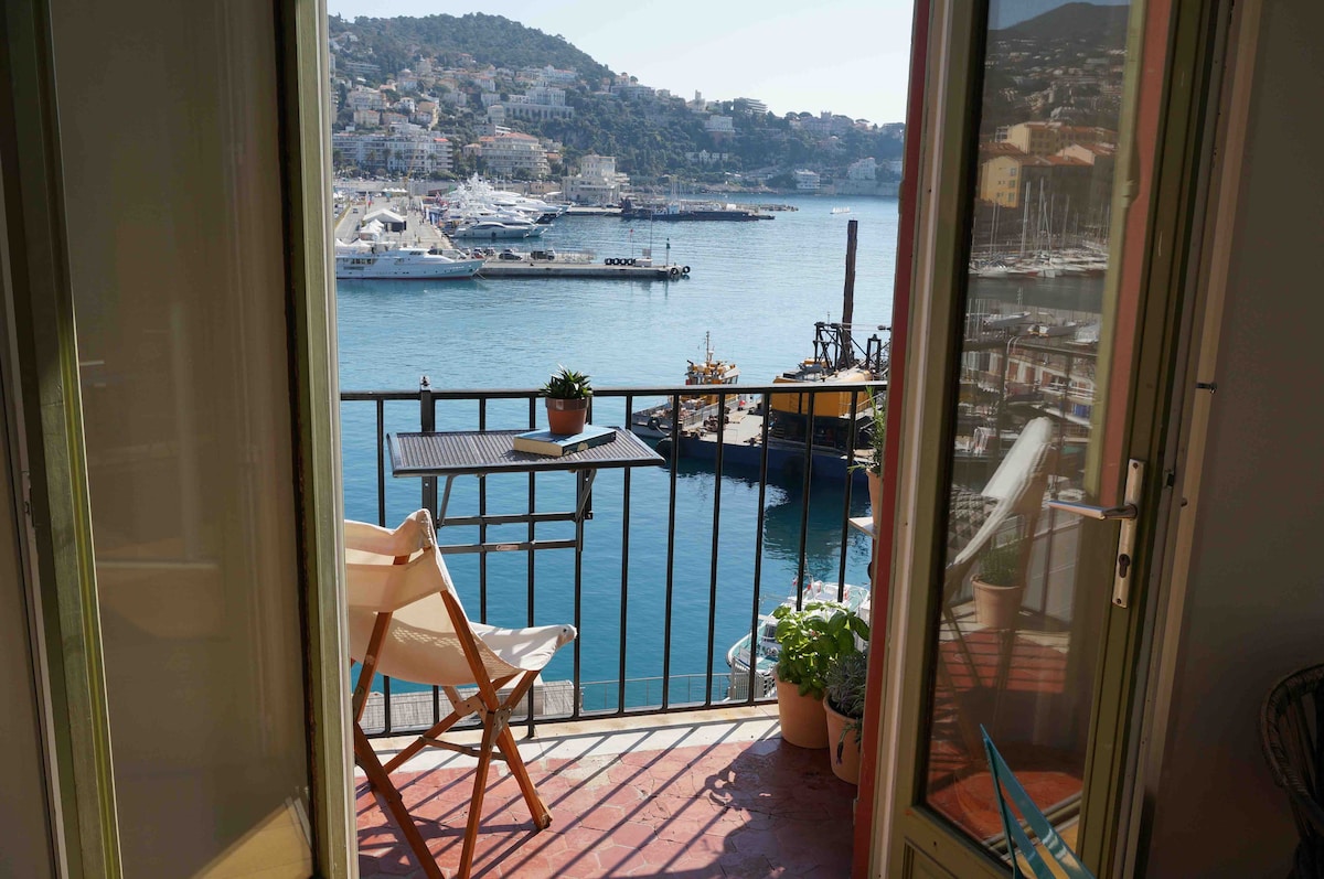 A sunny balcony offers a captivating view of the Mediterranean Sea and the port. A small table and a chair are placed on the balcony, surrounded by plants. The glass doors open to invite natural light and the beauty of the waterfront inside.