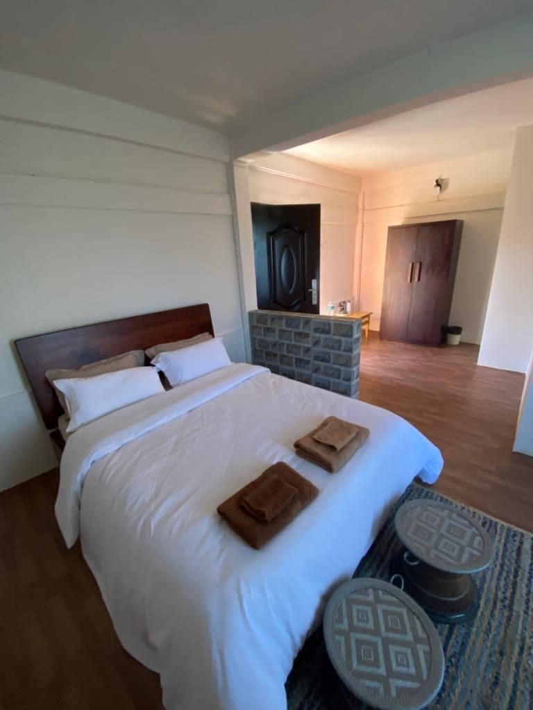 A spacious sleeping area features a wooden bed with crisp white linens. Soft towels are neatly arranged on the bed. Two small, patterned tables are positioned alongside. The room includes an open doorway leading to a cozy sitting area with a wooden table and chairs.