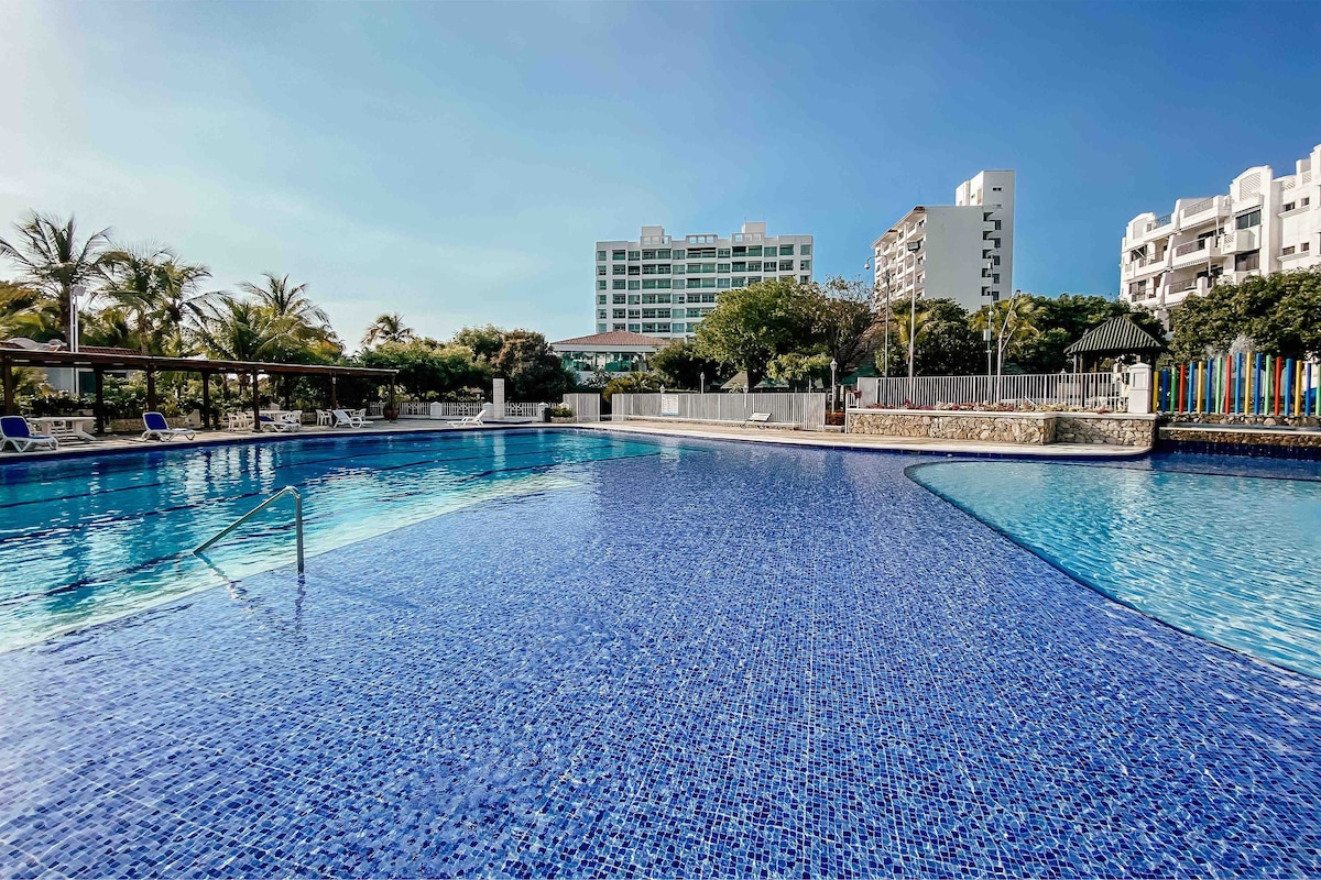 A large outdoor pool features clear blue water, with a vibrant tiled surface reflecting sunlight. Surrounding palm trees provide shade, and lounge chairs are positioned along the pool's edge. Nearby buildings and greenery create a serene environment for relaxation.