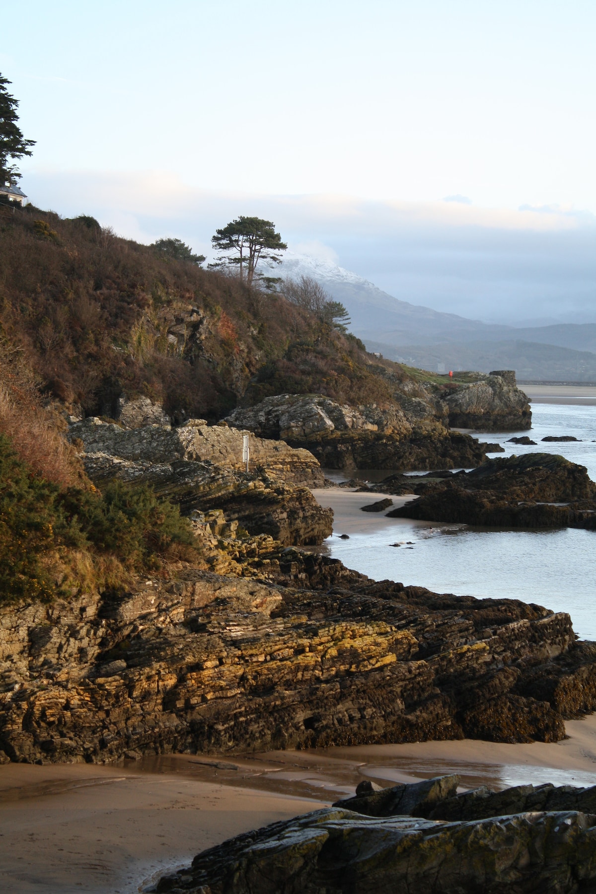 A rugged coastline displays distinct rock formations and sandy patches, bordered by gently sloping hills. Trees are scattered along the cliffs, providing a natural backdrop, while the calm water reflects the soft hues of the sky. The image captures the peaceful essence of the coastal landscape.