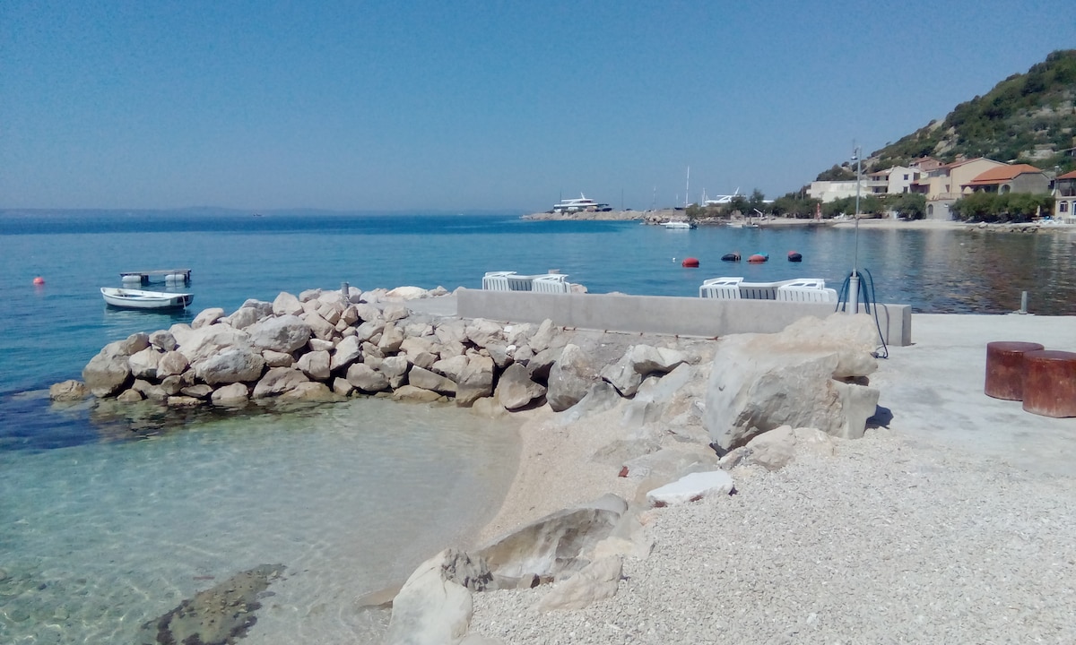 A calm coastline scene features clear blue water gently lapping against a rocky shore. A small boat is visible in the distance, while several moorings are positioned in the water. Sunlight reflects off the surface, enhancing the tranquil atmosphere.