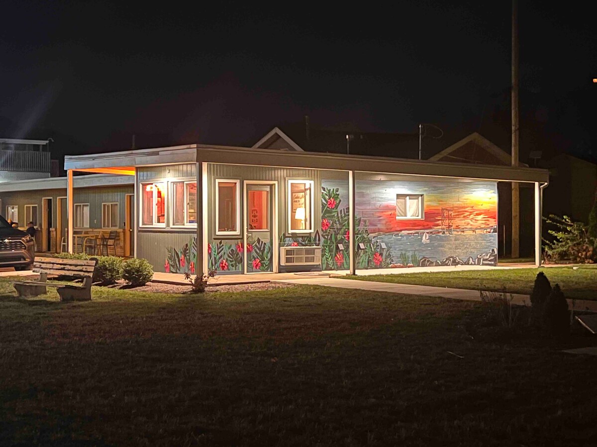 The exterior of a single-story motel is illuminated at night, highlighting a mural featuring a sunset over a water scene. Bright windows offer a glimpse of the interior. A park bench sits on the grassy front, enhancing the inviting atmosphere of the setting.