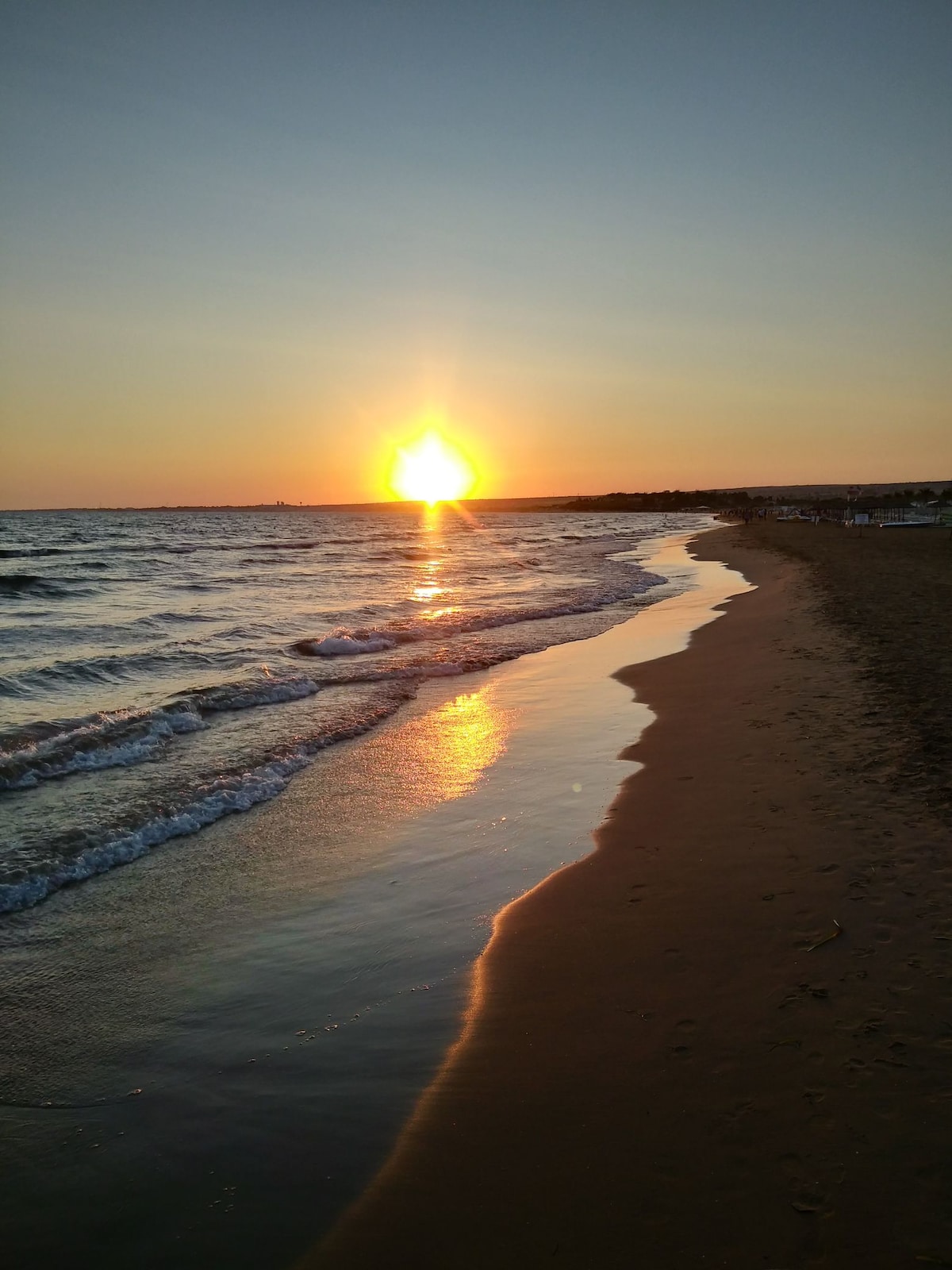 A beach is seen as the sun sets on the horizon, casting warm hues across the sky. Gentle waves lap at the shore, creating a reflective surface that captures the light. The sandy beach curves gracefully, inviting evening strolls along the coastline.