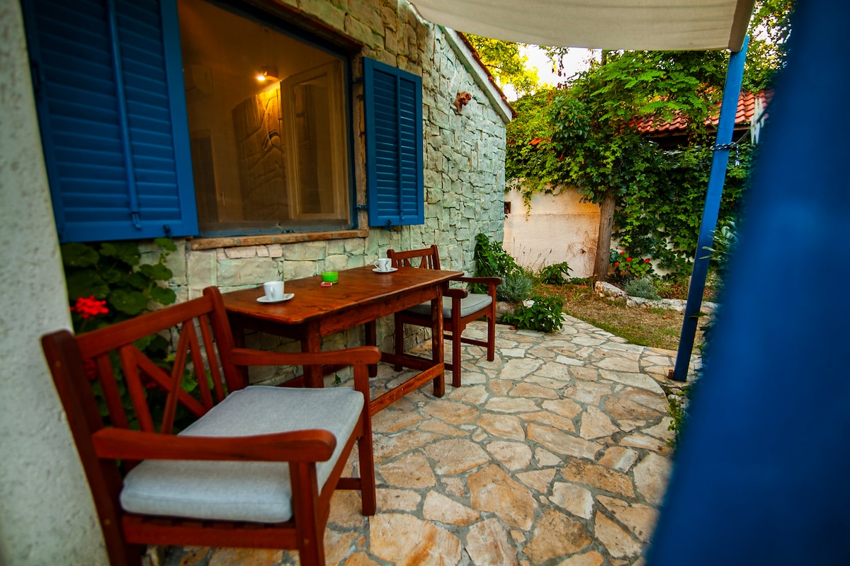 A small terrace features a wooden table and two chairs, surrounded by vibrant greenery and floral accents. Blue shutters frame the window, while a stone path leads through the lush yard, inviting relaxation in a peaceful outdoor setting.