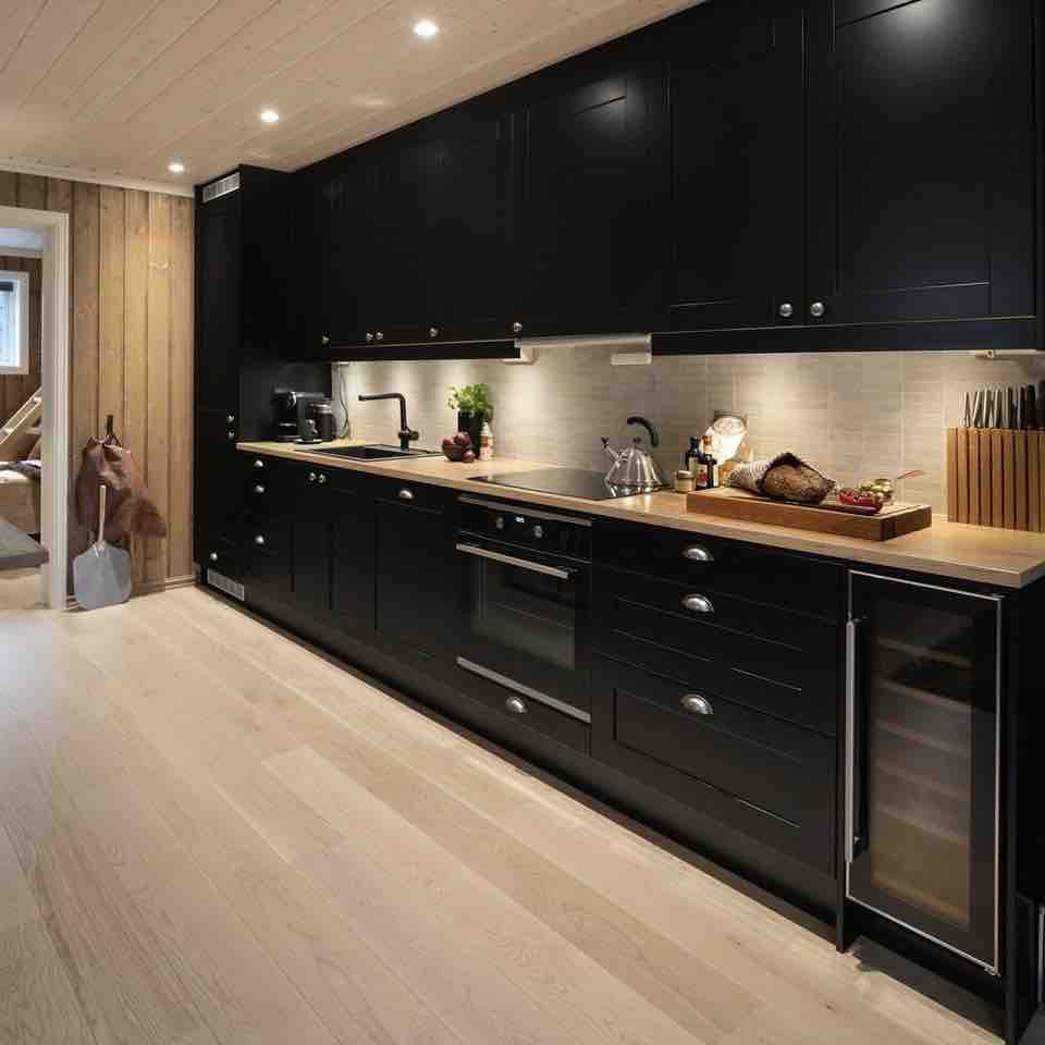 A modern kitchen features sleek black cabinetry with a wooden countertop. A built-in wine refrigerator is visible, along with a stove and a sink. Various kitchen essentials are arranged on the counter, complemented by soft overhead lighting that highlights the clean design.