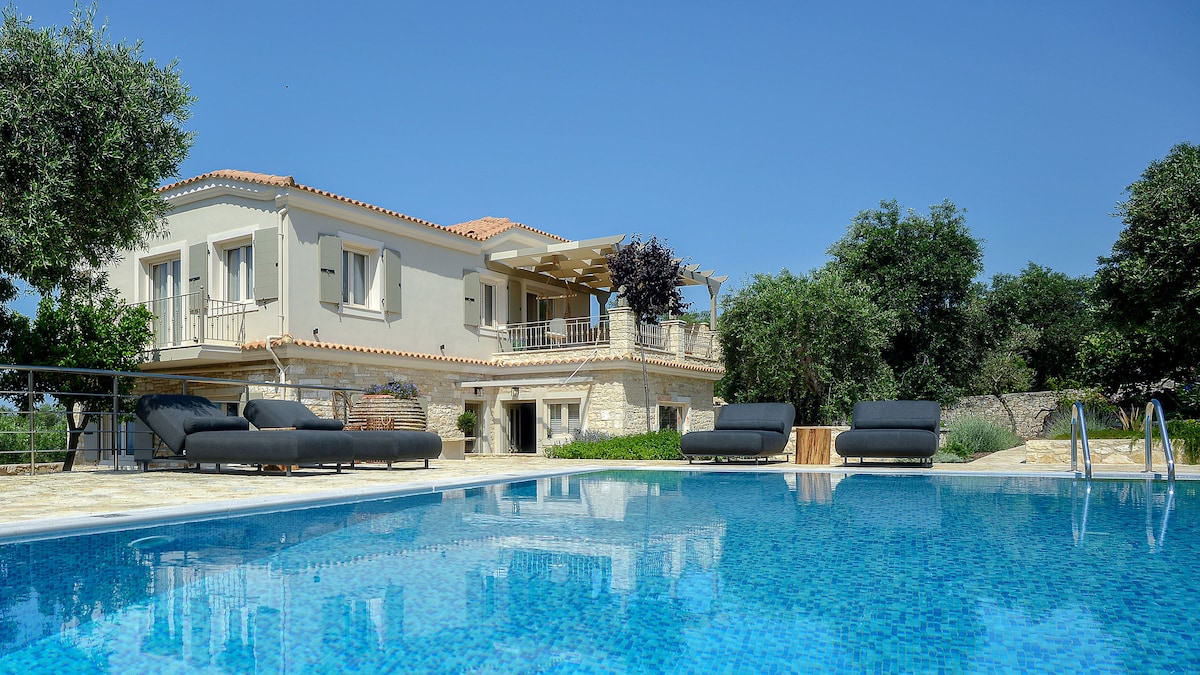 The spacious villa is showcased with its elegant design and balconies. A well-maintained swimming pool reflects the clear blue sky, while surrounding olive trees provide natural shade. Comfortable lounge chairs are placed poolside, offering a relaxing space to enjoy the serene outdoor setting.