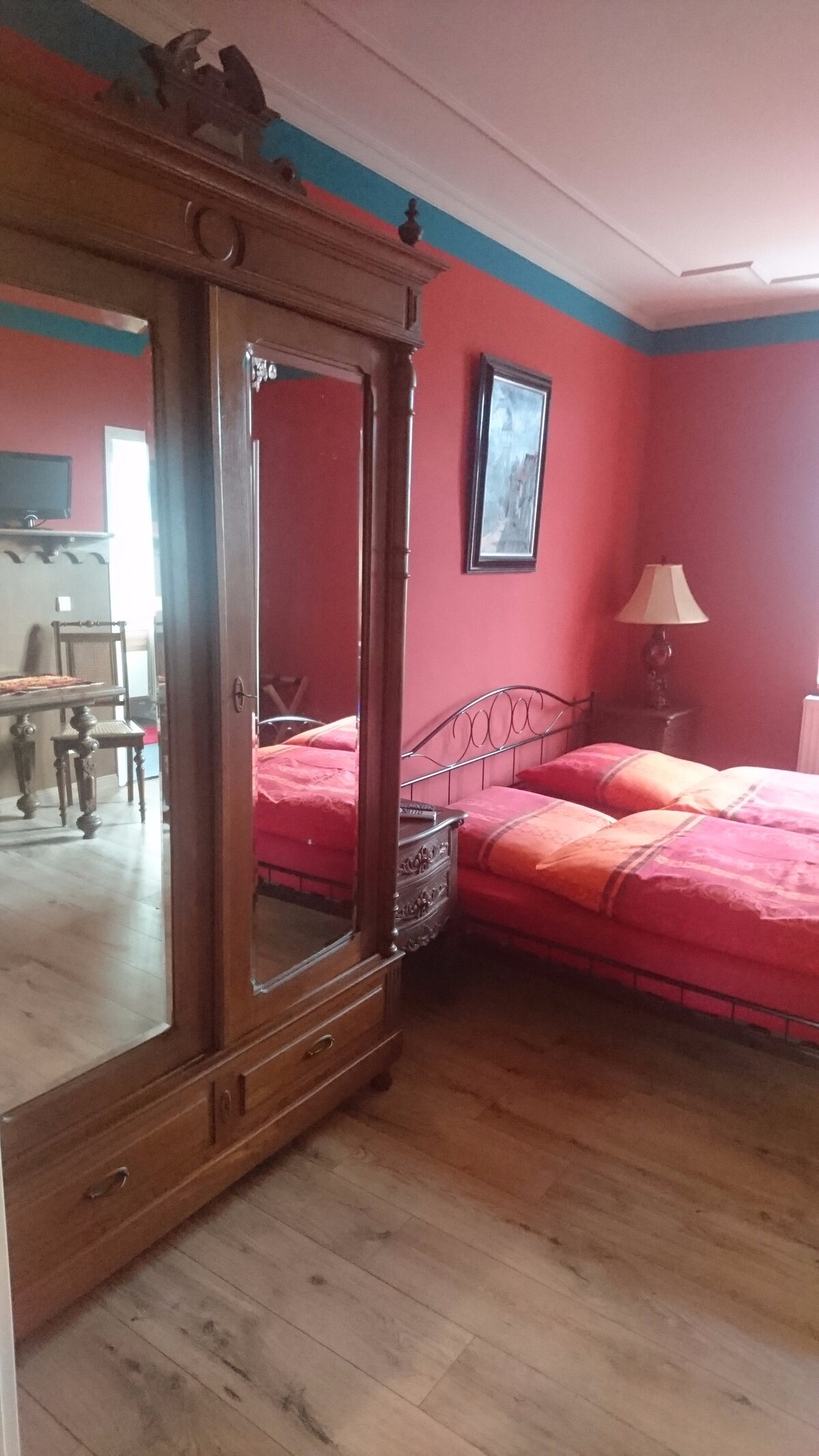 A cozy room features a spacious bed with bright red bedding, flanked by a decorative wooden wardrobe reflecting the room's design. A bedside lamp provides soft lighting, while warm wood flooring complements the vibrant red walls.
