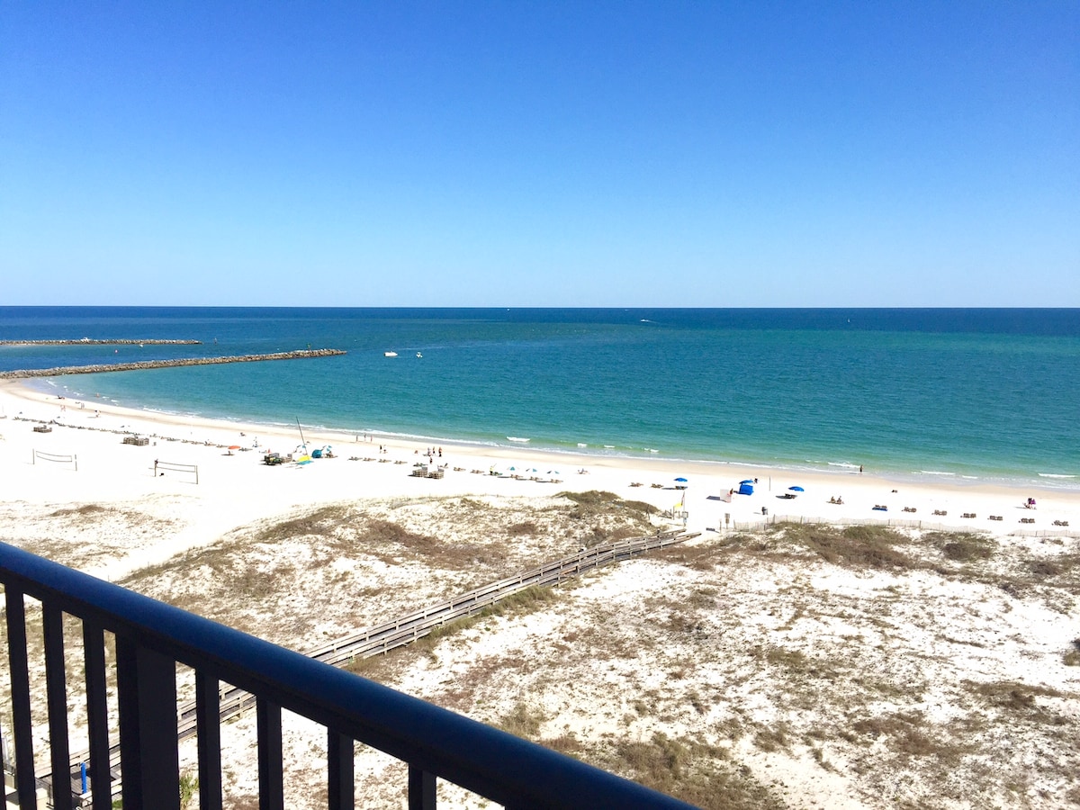 A panoramic view from the sixth-floor balcony showcases a serene beach with soft, sandy expanses. Gentle waves lap against the shoreline, while a few distant boats are visible on the horizon. The clear blue sky complements the vibrant colors of the Gulf waters.