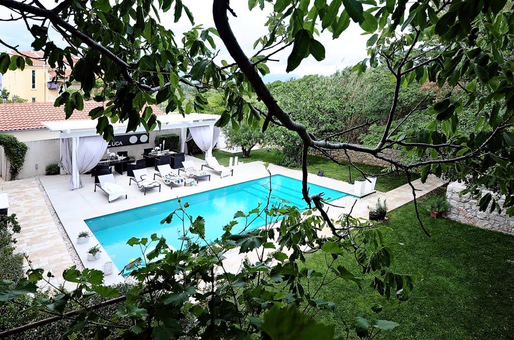 A serene swimming pool is surrounded by a spacious patio, furnished with lounge chairs and shaded areas. Lush greenery frames the view, including climbing vines and trees, creating a calm, private atmosphere. The inviting pool reflects the surrounding nature, providing a tranquil retreat.