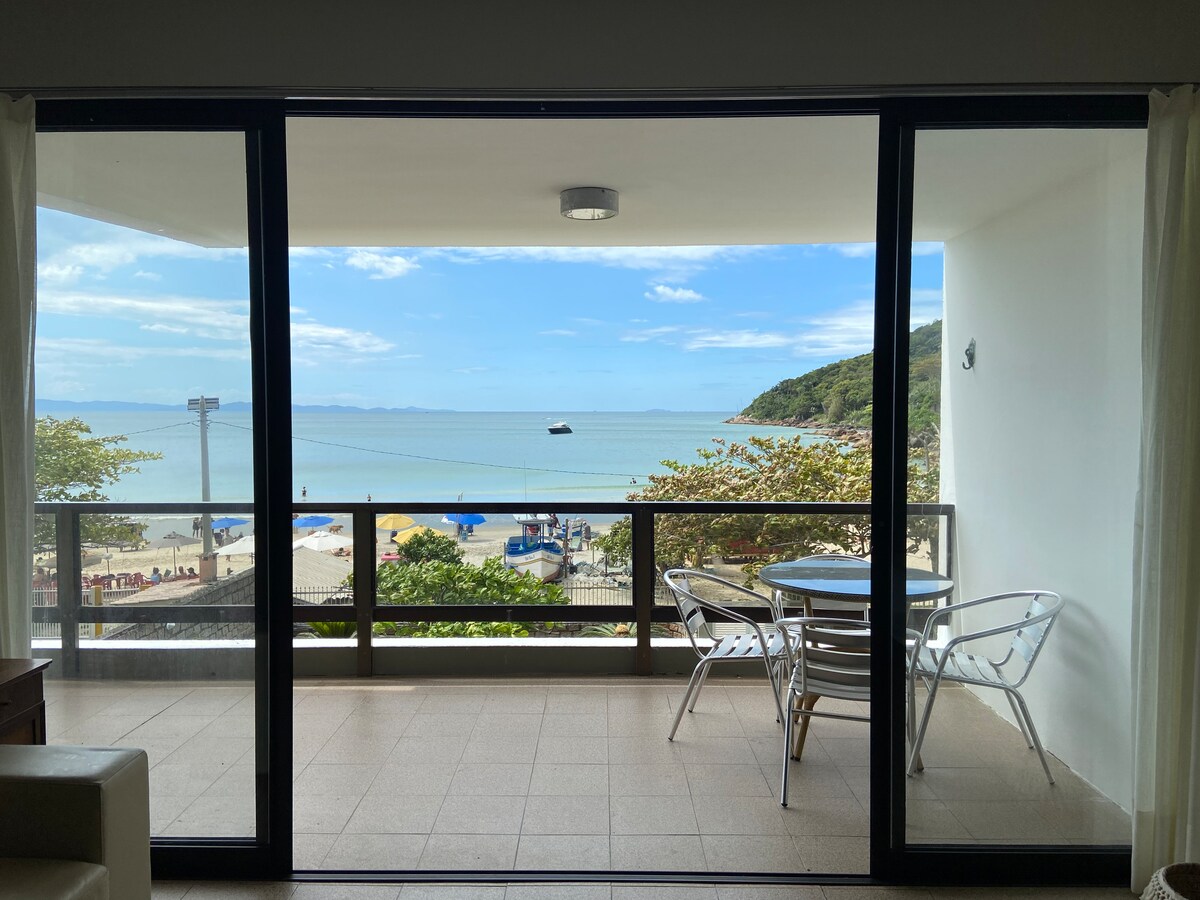 A spacious balcony is visible through floor-to-ceiling glass doors, offering a clear view of the calm ocean and beach. A small round table with metal chairs is positioned on the balcony, inviting relaxation with the gentle sound of the waves.
