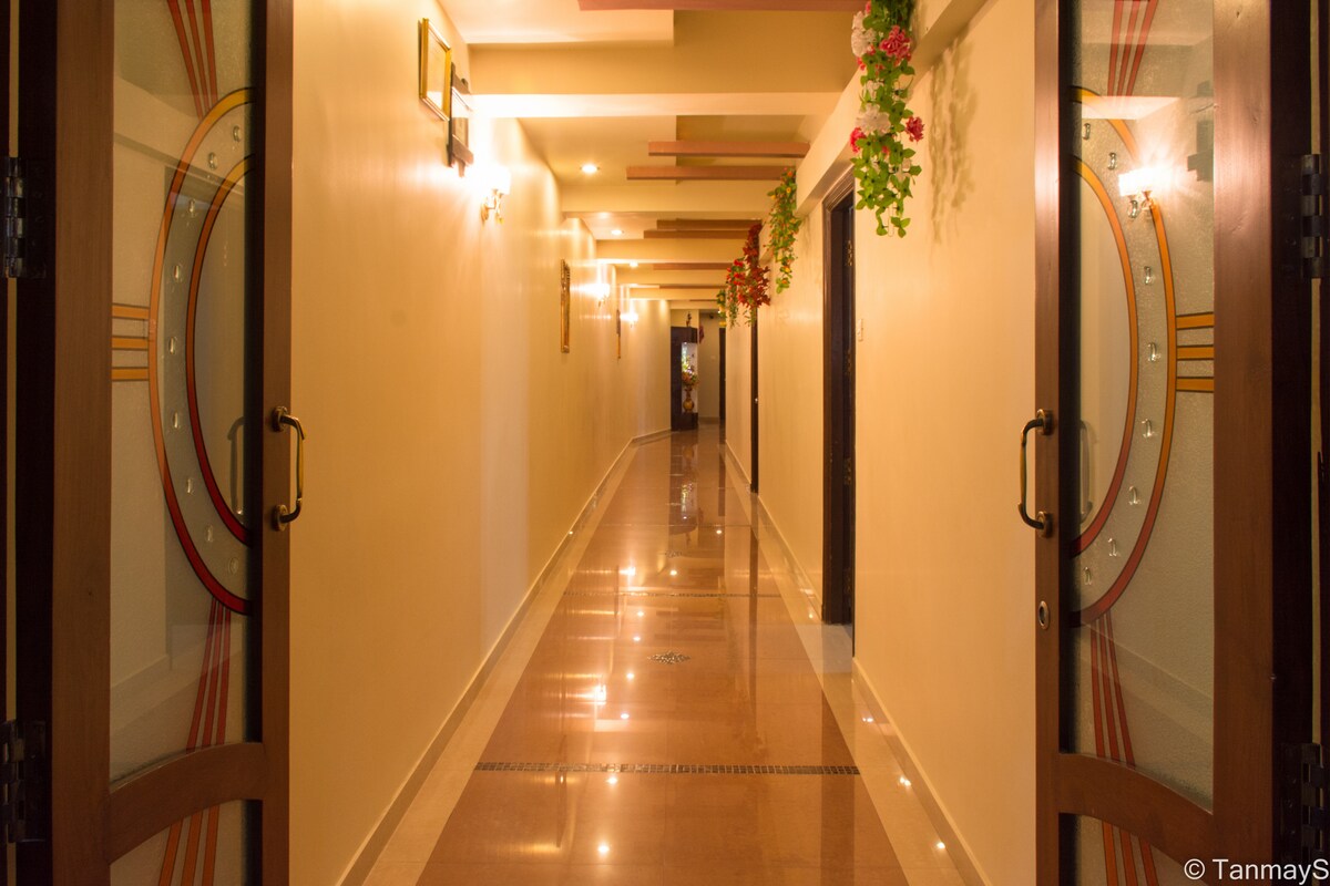 A well-lit corridor features polished tile flooring and softly glowing wall sconces. Decorative plants hang from the ceiling, adding a touch of greenery. Mirrors are placed on either side of the entrance, reflecting the warm lights and creating a welcoming atmosphere.