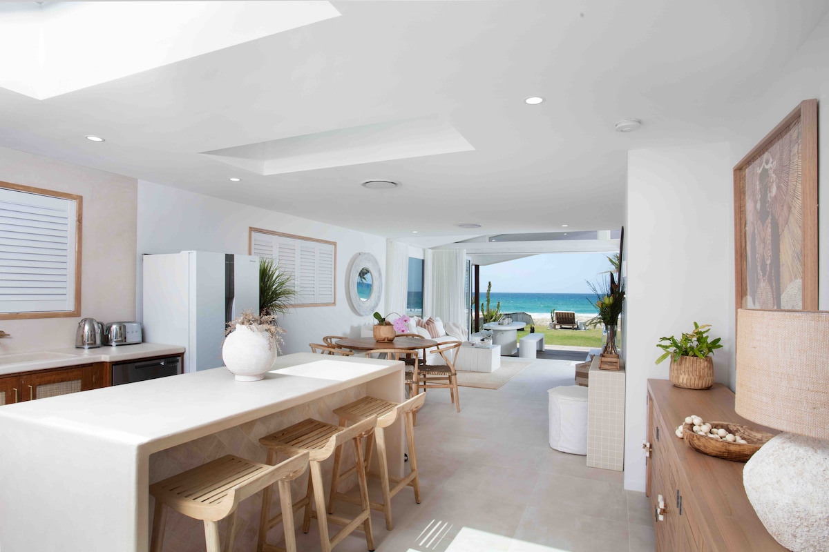 An open-concept living area showcases a modern kitchen with a large island and seating. A dining table is set nearby, while the expansive glass doors provide unobstructed views of the beach and ocean. Natural light highlights the neutral color palette throughout the space.